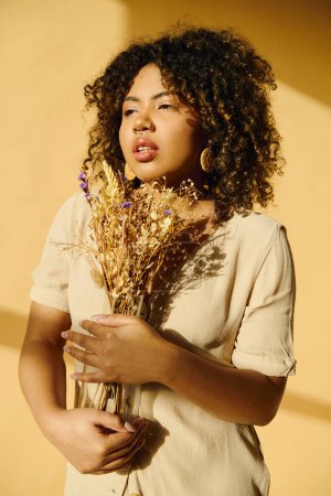 Foto de Una hermosa joven afroamericana con el pelo rizado en un vestido de verano delicadamente sosteniendo un ramo de flores secas. - Imagen libre de derechos