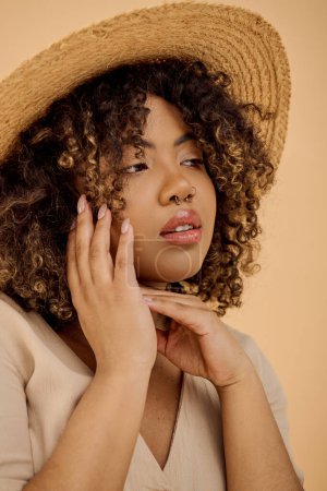 A beautiful young African American woman with curly hair holds her hand to her face, wearing a straw hat and a summer dress.