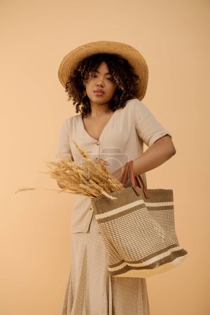 Joven mujer afroamericana con el pelo rizado en un sombrero de paja con elegancia sosteniendo una bolsa en un entorno de estudio brillante.