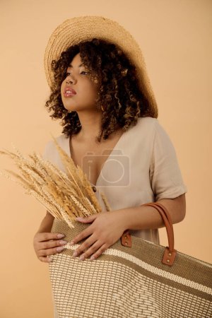 Téléchargez les photos : Jeune femme afro-américaine aux cheveux bouclés, portant un chapeau de paille, tenant un sac dans un cadre élégant studio. - en image libre de droit