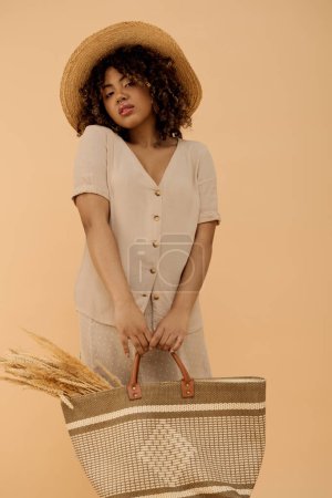 Téléchargez les photos : Jeune femme afro-américaine aux cheveux bouclés, dans un chapeau et une robe d'été, tenant un élégant sac de paille dans un cadre de studio serein. - en image libre de droit