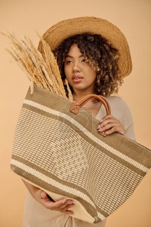 Foto de Hermosa mujer afroamericana con el pelo rizado en un vestido de verano sosteniendo una bolsa con un sombrero de paja en la parte superior en un entorno de estudio. - Imagen libre de derechos