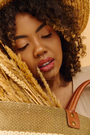 Eine schöne junge Afroamerikanerin mit lockigem Haar hält elegant eine braune Tasche in der Hand, während sie im Studio einen Strohhut trägt.