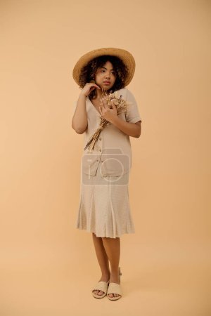Foto de Una joven y elegante mujer afroamericana con el pelo rizado posa con confianza en un vestido de verano y sombrero para un retrato. - Imagen libre de derechos