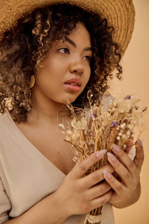 Foto de Una hermosa joven afroamericana con el pelo rizado, vestida con un vestido de verano, sosteniendo un ramo de flores secas. - Imagen libre de derechos