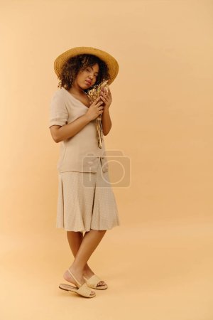 Eine schöne junge Afroamerikanerin mit lockigem Haar hält eine Blume in der Hand, während sie in einem Studio einen stylischen Hut trägt.