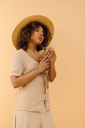A beautiful young African American woman with curly hair wearing a straw hat, holding dried flowers in a peaceful pose.