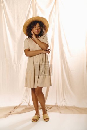 A beautiful young African American woman with curly hair strikes a pose in a summer dress and hat in a studio setting.