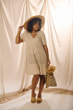 A beautiful young African American woman with curly hair poses gracefully in a stylish dress and hat in a studio setting.