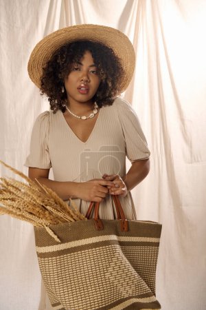 Téléchargez les photos : Une femme afro-américaine élégante aux cheveux bouclés, portant un chapeau de paille et tenant un sac dans un cadre de studio. - en image libre de droit