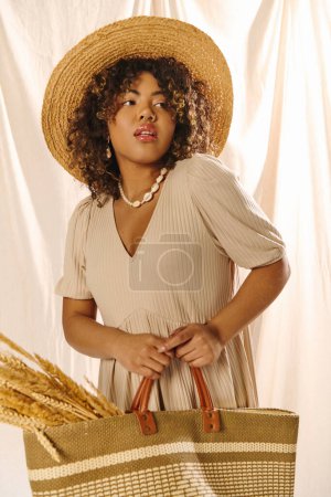 Eine junge Afroamerikanerin mit lockigem Haar posiert elegant mit einem Strohhut, während sie im Studio einen Strohsack in der Hand hält.