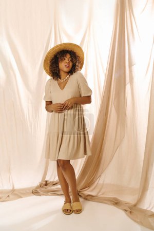 Eine elegante junge Afroamerikanerin mit lockigem Haar und Hut steht selbstbewusst vor weißem Hintergrund in einem Atelier.