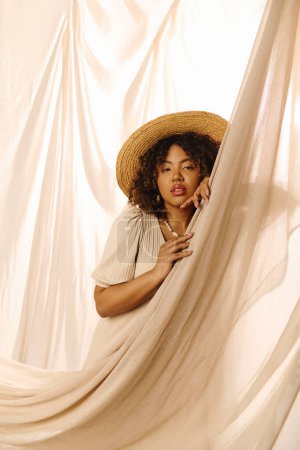 Eine schöne junge Afroamerikanerin mit lockigem Haar und Strohhut guckt aus einem Vorhang in einem Studio-Setting.