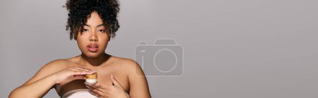 African American woman with curly hair holding a jar of cream in a studio setting, showcasing skin care and beauty routine.