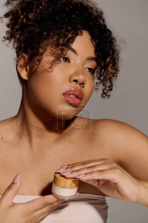 A beautiful young African American woman with curly hair tenderly holds a jar of cream in a studio setting.