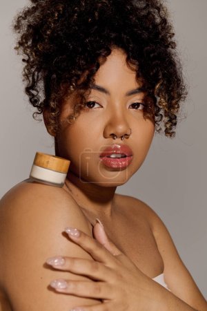 A young African American woman with curly hair elegantly balances a jar of cream on her shoulder in a studio setting.