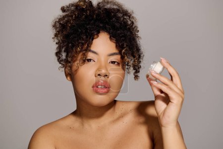 Foto de Joven mujer afroamericana con pelo rizado sosteniendo tarro de crema facial - Imagen libre de derechos