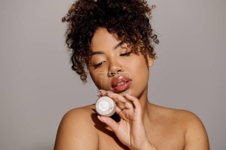 Young African American woman with curly hair holding jar of cream to her face for skin care routine in a studio setting.
