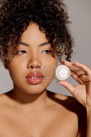 A young African American woman with curly hair holds a jar of cream in front of her face, emphasizing skincare and beauty.