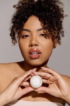 Téléchargez les photos : Une belle femme afro-américaine aux cheveux bouclés tenant délicatement un pot de crème dans ses mains. - en image libre de droit