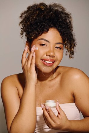 Beautiful African American woman with curly hair holding a jar of cream in front of her face in a studio setting.