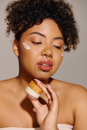 Young African American woman with curly hair applying cream from a jar onto her face in a studio setting.
