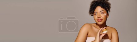 Photo for A beautiful young African American woman with curly hair, wrapped in a towel, happily holds a cream jar - Royalty Free Image