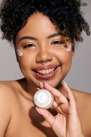 Young African American woman with curly hair applying cream from a jar to her face in a studio setting.