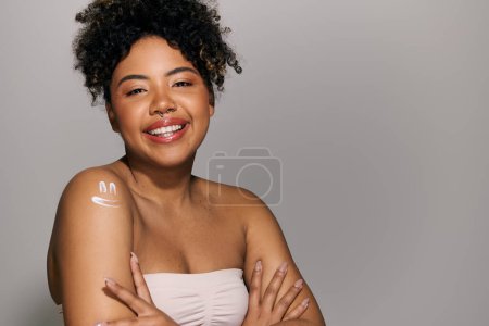 A beautiful young African American woman with curly hair poses confidently, crossing her arms for a portrait in a studio setting.