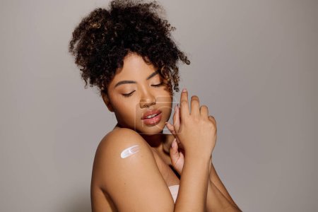 Foto de A beautiful young African American woman with curly hair pampering her skin with a white cream in a studio setting. - Imagen libre de derechos