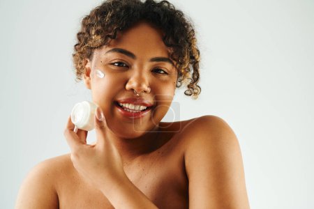 Photo for Smiling African American woman holds cream near face. - Royalty Free Image