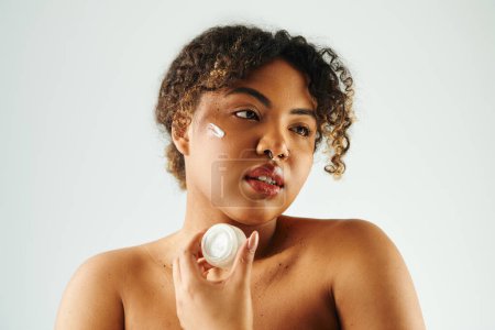 Shirtless African American woman gracefully holds a bottle of cream.