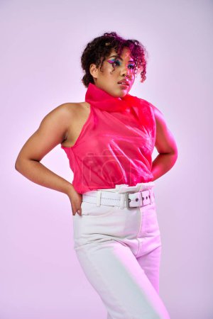 Beautiful African American woman poses in pink shirt and white pants on vibrant backdrop.