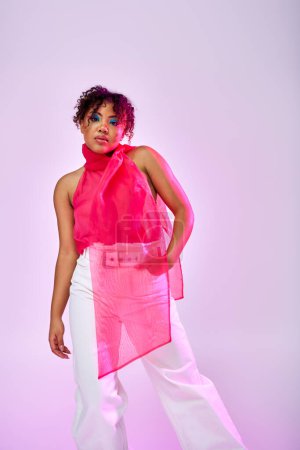 African American woman posing gracefully in pink top and white pants on vibrant backdrop.