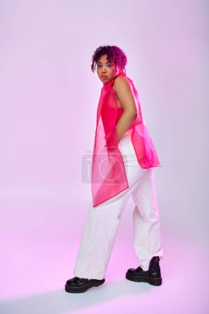 Una hermosa mujer afroamericana posa activamente en un top rosa y pantalones blancos sobre un vibrante telón de fondo.