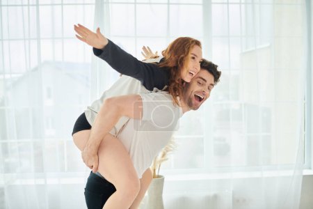 Foto de Un hombre sin esfuerzo levanta a una mujer sobre su espalda, mostrando fuerza y amor en un hermoso entorno de dormitorio. - Imagen libre de derechos