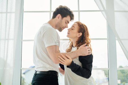 Téléchargez les photos : Un homme et une femme se tiennent tranquillement côte à côte devant une fenêtre, embrassant un moment serein ensemble. - en image libre de droit