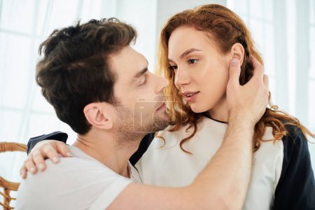 Foto de Un hombre besa tiernamente a una mujer en la mejilla en un ambiente de dormitorio, mostrando amor e intimidad entre ellos. - Imagen libre de derechos