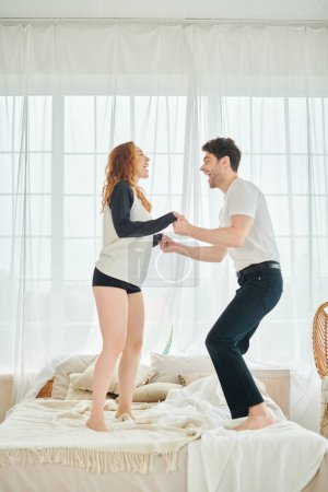 A man and woman stand atop a bed, sharing a moment of intimacy and connection in a cozy bedroom setting.