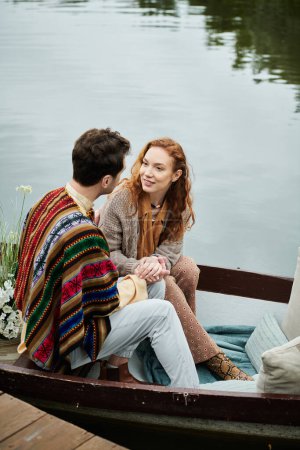 A man and a woman, dressed in boho style clothes, sit peacefully in a boat surrounded by lush greenery on a romantic date in a park.