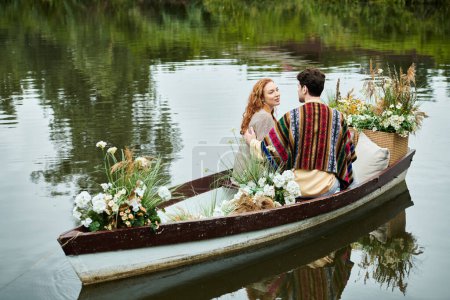 Ein Mann und eine Frau in Boho-Kleidung rudern in einem Boot voller lebendiger Blumen in einem üppig grünen Park und genießen ein romantisches Date.