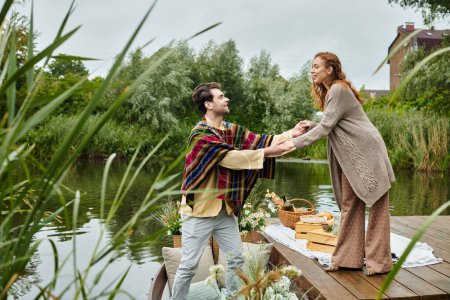 A couple in boho attire stands hand in hand on a wooden dock, surrounded by greenery and calm waters.