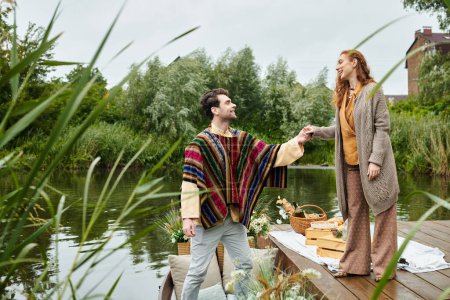 A man and a woman in boho style clothes stand on a dock, enjoying a serene moment by the water in a green park setting.