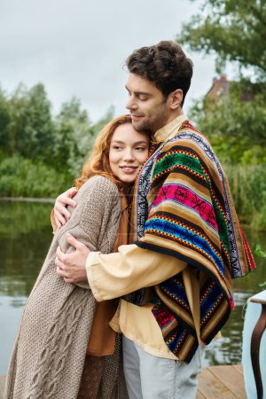 A man and woman in boho attire stand beside a tranquil body of water in a beautiful park setting.