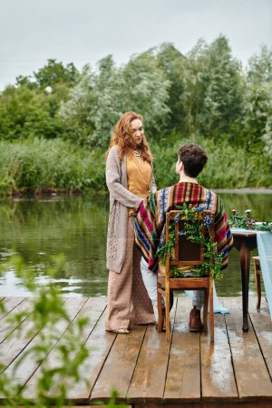 A romantic couple in boho style attire embrace while sitting on a picturesque dock in a serene green park setting.