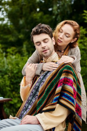 A man tenderly holds a woman wrapped in a blanket, their boho style clothes blending into the lush green backdrop of a park.