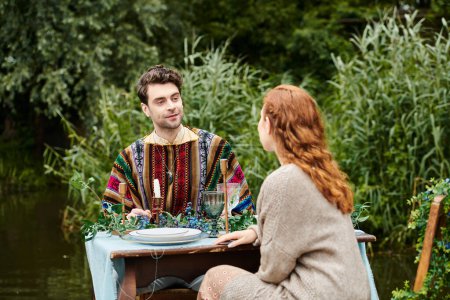 A man and a woman in boho style clothing sit together at a table in a green park, enjoying a romantic date by a tranquil pond.