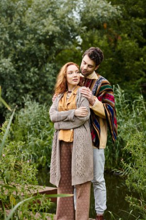 Téléchargez les photos : Un homme et une femme en tenue boho se tiennent côte à côte sur un pont en bois dans un parc verdoyant, partageant un moment tendre. - en image libre de droit