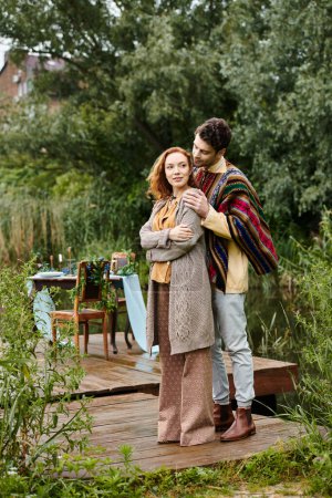 Foto de Un hombre y una mujer, vestidos con ropa de estilo boho, están en un muelle junto a un lago sereno. - Imagen libre de derechos