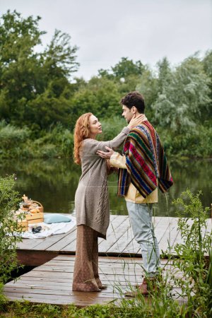 A man and a woman, stylishly dressed in boho attire, stand together on a peaceful dock in a lush green park.